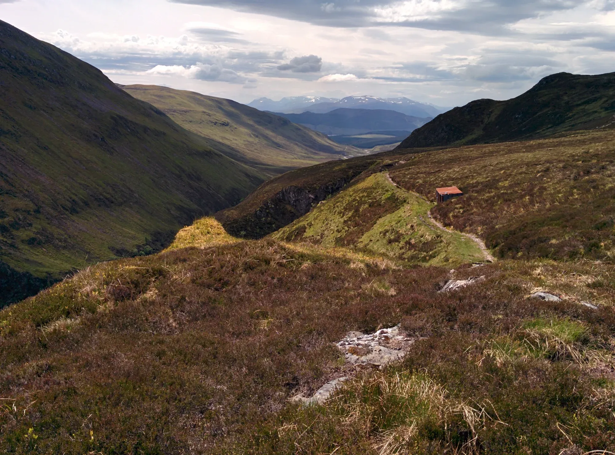 Mountain path across heather moorland — a small bothy visible in the distance, long valley views and dramatic clouds