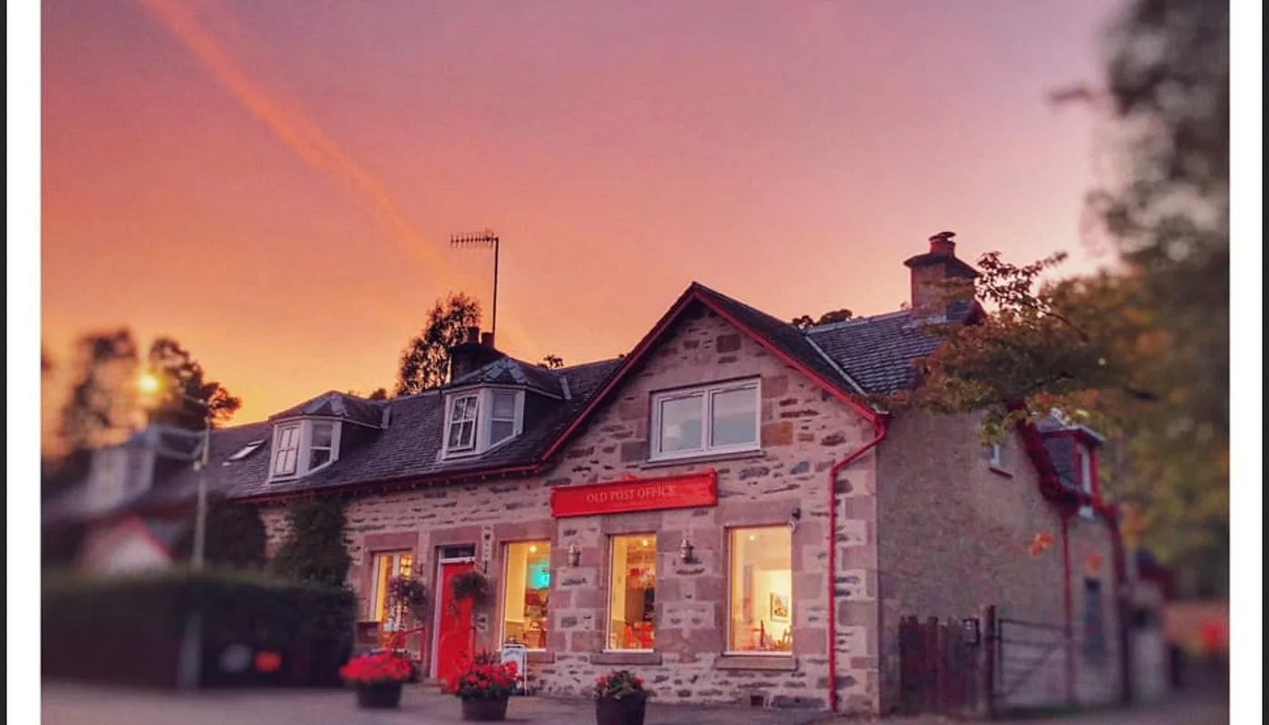 Stone cottage with a red Old Post Office sign, red door and lit windows under a pink and orange sunset sky.